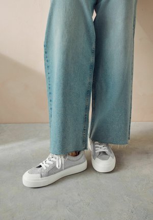 Sneakers grigi con tessuto texture e suole bianche, abbinati a jeans a gamba larga blu chiaro con orli sfrangiati.