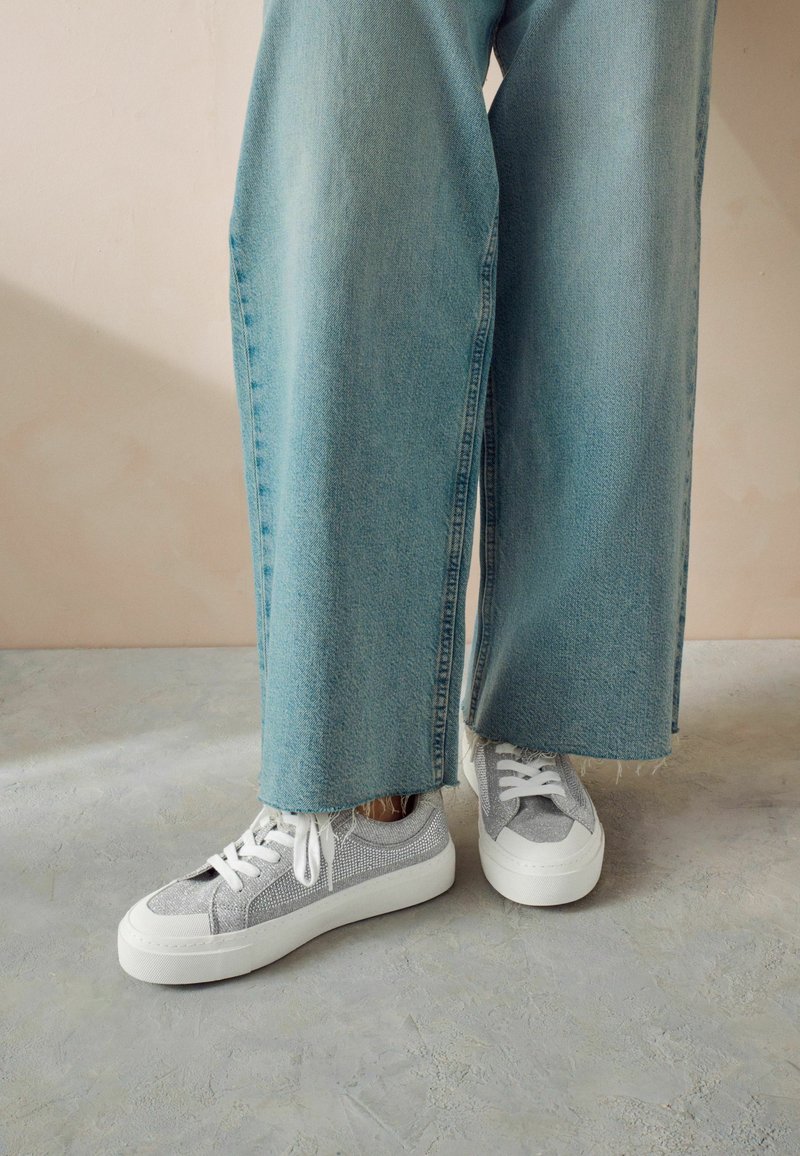 Sneakers grigi con tessuto texture e suole bianche, abbinati a jeans a gamba larga blu chiaro con orli sfrangiati.
