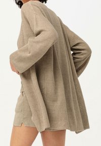 Beige stickad cardigan med långa ärmar, ribbad struktur och sidspringor. Har en avslappnad passform och når ner under höften.