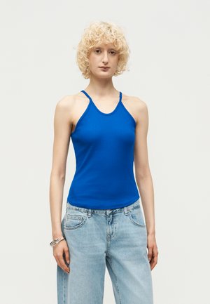 Personne aux cheveux blonds bouclés portant un haut sans manches bleu vif et un jean bleu clair, debout devant un fond clair uni.