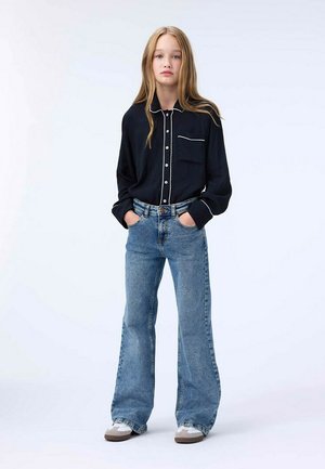 Camicia nera con bottoni e piping bianco, abbinata a jeans blu a vita alta e scarpe da ginnastica chiare. Maniche a taglio dritto e taschino sul petto.