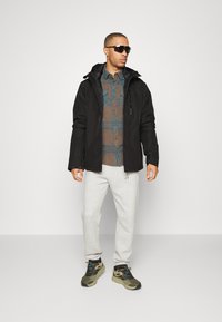 Chaqueta impermeable negra, camisa a cuadros marrón y azul, pantalones de chándal grises y zapatillas verde oliva. Los accesorios incluyen gafas de sol.