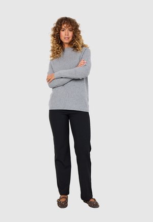 Gray ribbed sweater with a round neck, paired with black trousers, and brown flats adorned with gold studs. Model standing with arms crossed.