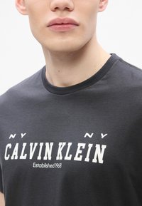 Hallisin halli puuvillane t-särk, lühikeste varrukatega, ümar kaelus, esiküljel suur valge tekst "CALVIN KLEIN" ja "Asutatud 1968".
