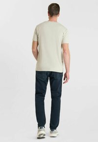 Uomo in piedi di spalle, che indossa una maglietta beige a maniche corte, jeans blu scuro e sneakers bianche e grigie su uno sfondo neutro.