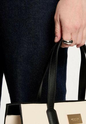 Mano con un anello d'argento che tiene una crema con manico nero e una borsa nera su sfondo di tessuto denim scuro.