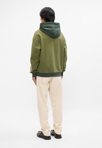 Ελιάς πράσινη hooded sweatshirt με αντίθετο σκούρο καπέλο, ριμπ μανσέτες και στρίφωμα, συνδυασμένη με ανοιχτό μπεζ παντελόνι και μαύρα παπούτσια.