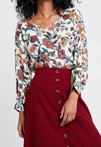 Bloemige blouse met multikleurige patronen op een crèmekleurige achtergrond, gecombineerd met een bordeauxrode rok met knopen aan de voorkant.