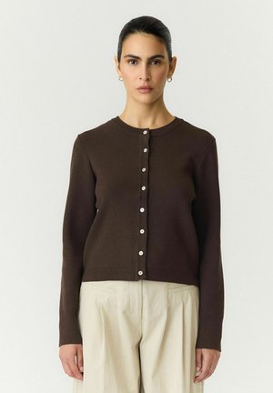 Femme aux cheveux foncés en chignon, portant un cardigan marron foncé boutonné et un pantalon beige clair, debout devant un fond clair uni.