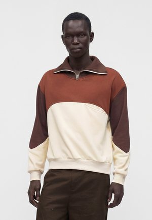 Hombre que lleva un jersey color block con secciones en marrón, crema y marrón oscuro, con un cuello con cremallera, de pie frente a un fondo liso.