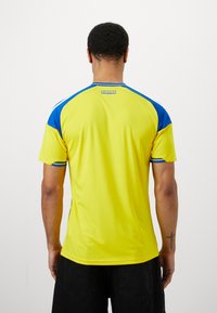 Man standing with back to camera wearing yellow and blue short-sleeve sports jersey with "Sverige" text and black shorts, plain white background.