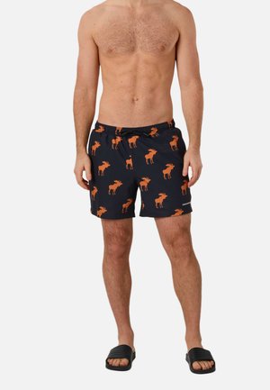 Mężczyzna w czarnych kąpielówkach z pomarańczowymi wzorami łosi i czarnych klapkach, stojący na jednolitym białym tle.