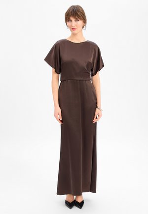 Femme debout portant une robe longue en satin marron à manches courtes et des escarpins noirs à bout pointu, sur fond blanc uni.