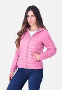 Donna con lunghi capelli castani che indossa una giacca rosa trapuntata, una camicia bianca e jeans blu, in piedi con una mano in tasca contro uno sfondo bianco.