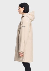 Bež jakna otporna na vodu s kapuljačom, featuring elastične manžete, bočne džepove i jednostavan dizajn. Logo otisnut na rukavu.