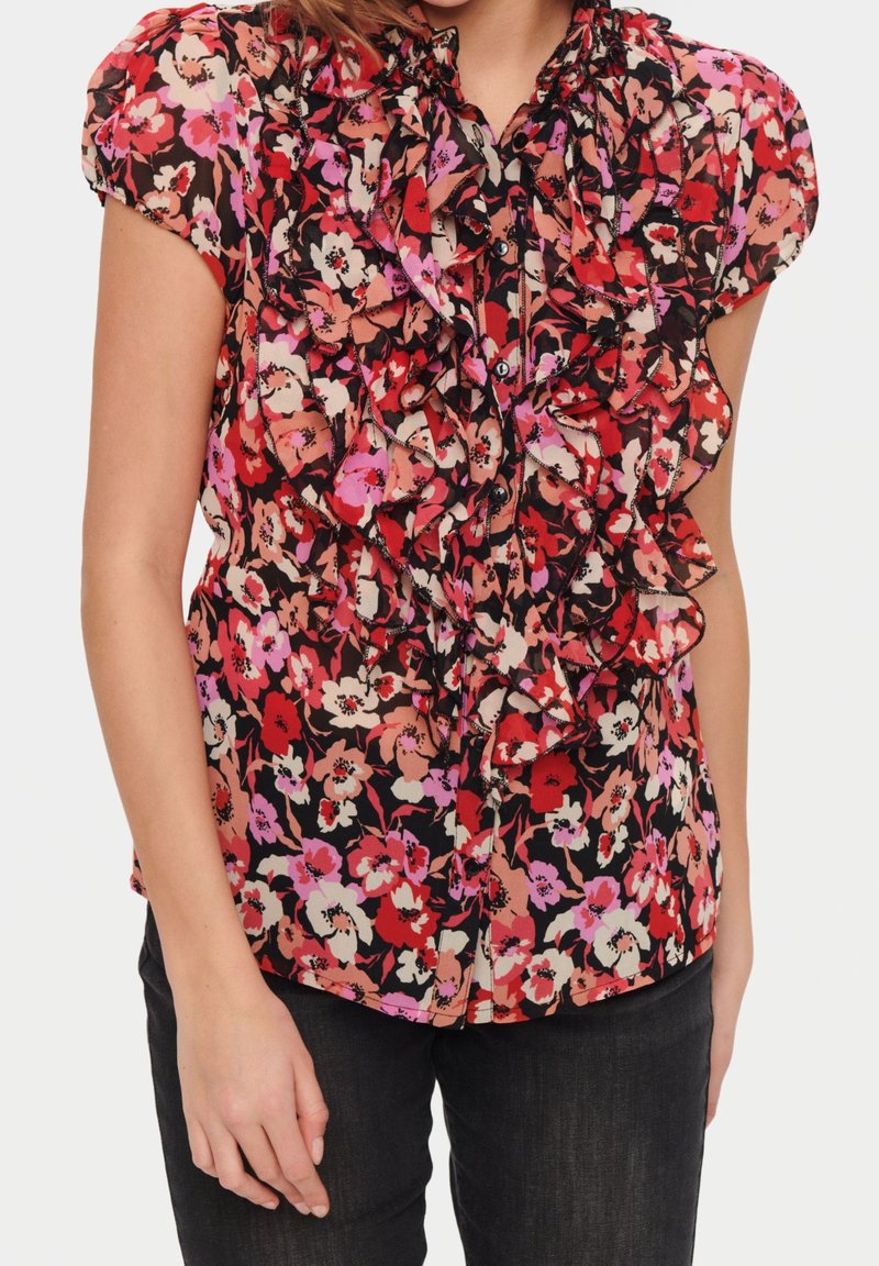 Femme portant un chemisier fleuri à manches courtes avec des volants rouges, roses et noirs, associé à un pantalon noir, sur fond blanc.