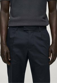 Námořnické kalhoty na míru s hladkou texturou, poutky na opasek, předními záhyby a bočními kapsami, v kombinaci s tmavým pleteným polo tričkem.