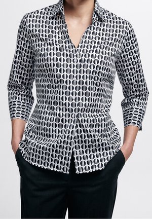 Frau trägt ein schwarz-weiß gemustertes Hemd mit Dreiviertelärmeln und schwarze Hose, Hände in den Taschen, vor weißem Hintergrund.