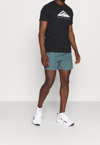 Camiseta corta de Nike Trail en negro, con gráfico de logotipo. Pantalones cortos deportivos verdes, de tejido ligero. Zapatillas deportivas blancas con detalles en contraste.