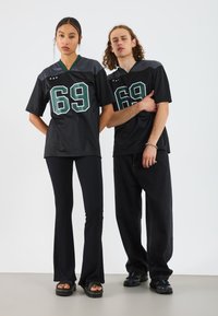 Svart sportsjersey med grønne tall "69," kombinert med svarte ribbede, utsvingte bukser. Modellen har på seg kraftige svarte sandaler. Minimalistisk design.