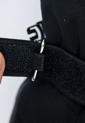 Cinturino in tessuto nero con superficie texturizzata, dotato di un anello in metallo argentato e di un piccolo dettaglio in pelle nera. Inquadratura ravvicinata della chiusura.