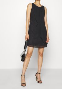 Robe noire sans manches avec un détail drapé à l'avant, tissu lisse et coupe sous le genou. Associée à des talons noirs à lanières et un petit sac à main.