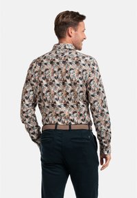 Chemise à motifs floraux avec manches longues, présentant des nuances de marron, bleu-vert et crème. Portée avec un pantalon sombre et une ceinture texturée.