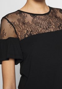 Svart kortärmad topp med volangärmar och genomskinlig spetskorsning som har blommönster och paisleymönster.