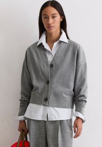 Cardigan en tricot gris avec un devant à boutons, des poignets côtelés et un ourlet. Superposé à une chemise rayée blanche et bleue. Le modèle tient un sac marron.