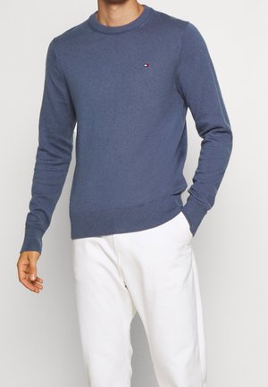 Homme portant un pull à col rond bleu à manches longues et un pantalon blanc, avec une main dans la poche et un petit logo sur la poitrine.
