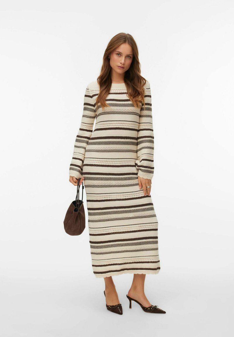 Stickad klänning med långärmade horisontella ränder i kräm och brunt, ribbad textur och knälång längd. Håller en brun väska.