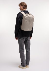 Zaino beige chiaro con chiusura a cordoncino, spallacci regolabili e una texture liscia, indossato da una persona in un ambiente neutro.