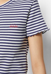 Námořnické a bílé pruhované tričko s vroubkovanou texturou, kulatým výstřihem a vlnkovými okraji rukávů. Obsahuje červený vyšitý text.