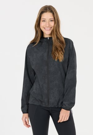 Junge Frau mit langen braunen Haaren lächelt und trägt eine schwarze Jacke mit Reißverschluss und schwarze Leggings vor einem schlichten hellen Hintergrund.