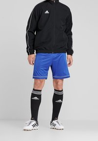 Svart zip-up jacka med vita ränder, blå shorts och svarta knästrumpor med vita ränder och logotyp, tillsammans med vita fotbollsskor.