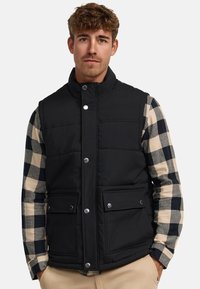 Chaleco acolchado negro con cuello alto, cierres de botón en la parte delantera y dos bolsillos. Lleva puesto una camisa de cuadros de manga larga en colores beige y azul marino.