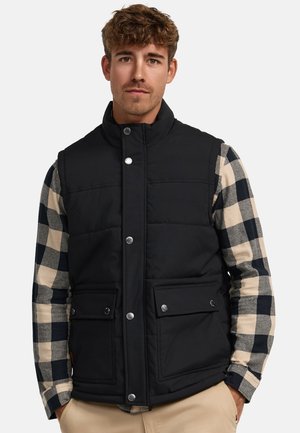 Black padded vest with high collar, front snap closures, and two pockets. Worn over a plaid long-sleeve shirt in beige and navy colors.