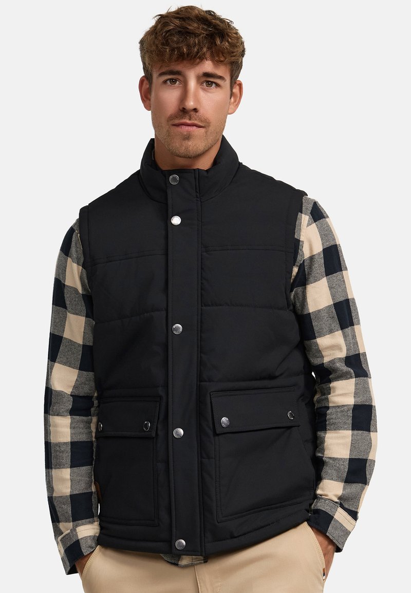 Chaleco acolchado negro con cuello alto, cierres de botón en la parte delantera y dos bolsillos. Lleva puesto una camisa de cuadros de manga larga en colores beige y azul marino.