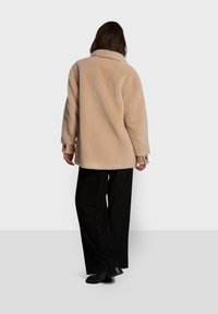 Casaco oversized de pelúcia beige com colarinho, mangas longas e textura suave, usado com calças largas pretas e sapatos pretos.
