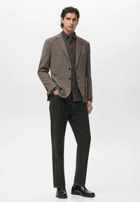 Blazer a motivo houndstooth marrone con due tasche frontali, abbinato a una camicia button-up scura e jeans scuri, completato da scarpe nere.