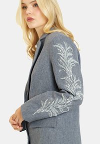 Blazer grigio con un design aderente, caratterizzato da motivi decorativi di foglie in paillettes argentate sulle maniche. Tessuto strutturato con una silhouette sartoriale.