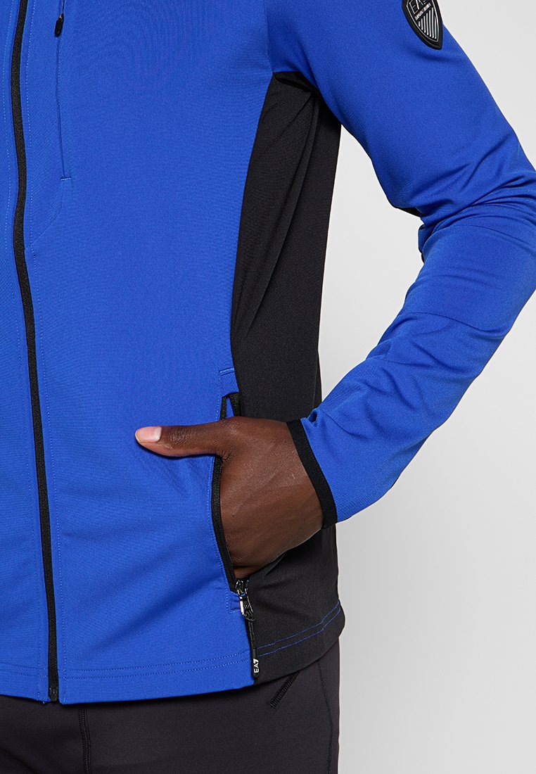 Giacca zip-up blu e nera con tessuto testurizzato, tasche laterali e patch del logo sulla spalla.