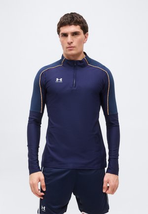 Mand iført mørkeblå Under Armour langærmet træningstrøje med kvartlynlås og matchende shorts, stående mod en ensfarvet baggrund.