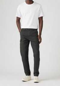 Cargo-Hosen aus dunkelgrünem Baumwollstoff mit mehreren Seitentaschen, gerader Beinform, kombiniert mit einem schlichten weißen Rundhals-T-Shirt und hellen Turnschuhen.
