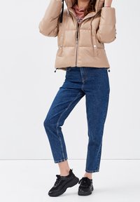 Veste matelassée beige avec fermeture éclair et boutons-pression, associée à un jean taille haute bleu et des chaussures de sport noires. Texture lisse, style court.