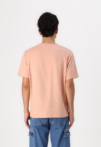 Tricou cu mânecă scurtă, guler rotund, din bumbac moale piersicuță, cu o croială lejeră și o textură fină. Vederea din spate arată detalii minime.