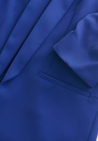 Blue blazer made of textured fabric. Features a notched collar, a single chest pocket, and folded sleeves, conveying a sleek design.