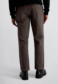 Homme portant un pantalon marron foncé avec des ourlets et des chaussures de ville noires, se tenant les mains détendues à ses côtés sur un fond clair.
