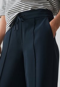 Marineblauwe cropped broeken met plooien, elastische tailleband en strikdetail. Ggedragen met een zwart-wit gestreept shirt met korte mouwen.