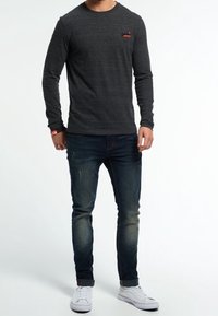 T-paita pitkällä hihalla, tummanharmaa, pehmeästä kankaasta, pyöreällä kaula-aukolla ja pienellä punaisella logolla vasemmassa rinnassa, yhdistettynä sinisiin farkkuihin ja valkoisiin lenkkareihin.